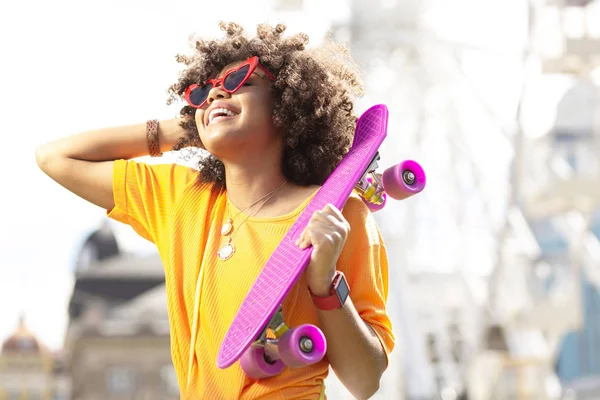 Веселая кудрявая девушка смеется, держа при себе мини скейтборд — стоковое фото