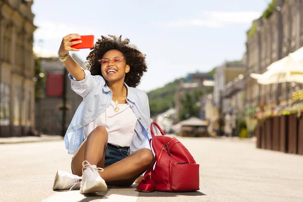 Beautiful moment. Upbeat young woman taking selfies and smiling at the camera while sitting on the asphalt in the streets