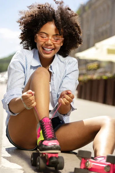 Upbeat lockigt tjejen ler medan kopplingsförbehåll roller skate skosnören — Stockfoto