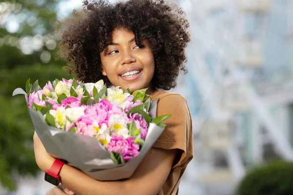 Best gift. Charming curly woman smiling widely and looking into the distance while holding a bouquet of flowers