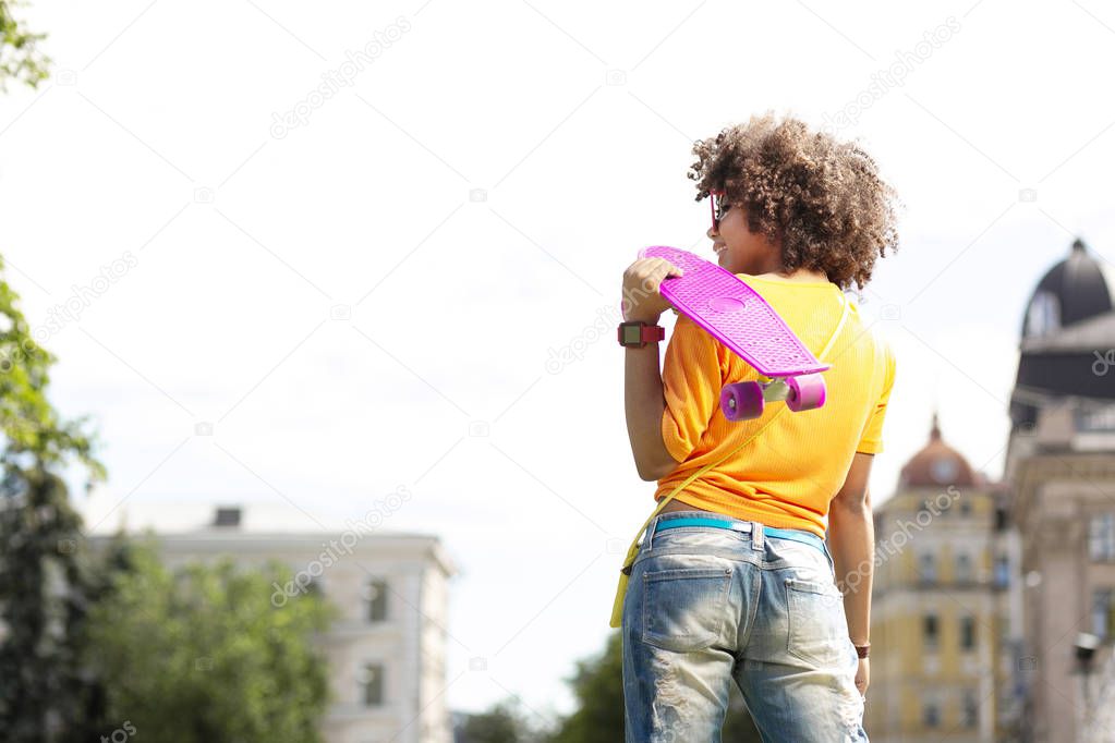 Cheerful curly woman standing with skateboard on shoulder