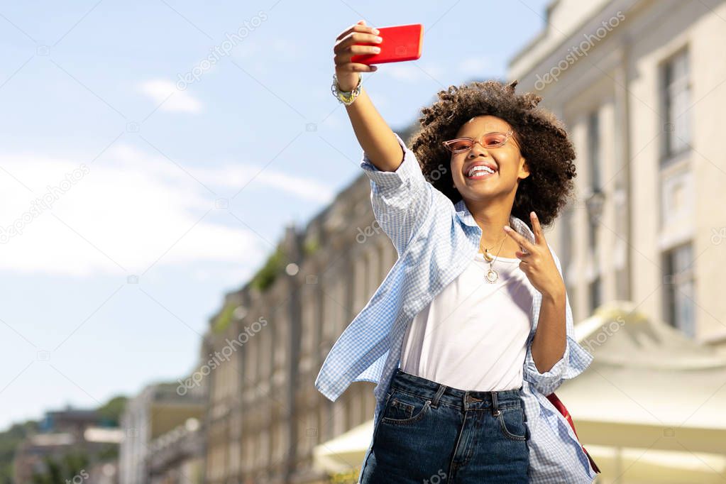 Joyful curly woman taking selfie and showing V sign