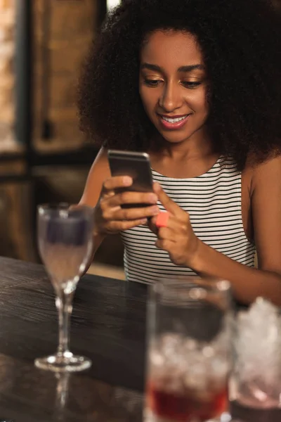 Beautiful curly woman texting while having cocktail in bar