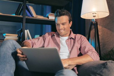 Evde. Koltukta oturan ve dizüstü bilgisayar ekranında bakarken gülümseyen mutlu güzel hoş adam