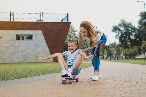 Ενθουσιασμένος αστείο κορίτσι που κάθεται στο skateboard κοντά τη μητέρα της — Φωτογραφία Αρχείου
