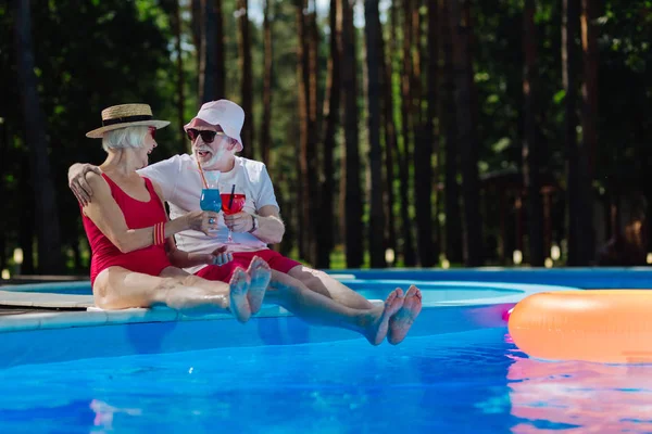 Couple of happy funny pensioners drinking cocktails near pool