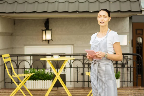 Прекрасна офіціантка стоїть на терасі кафе і тримає планшет — стокове фото