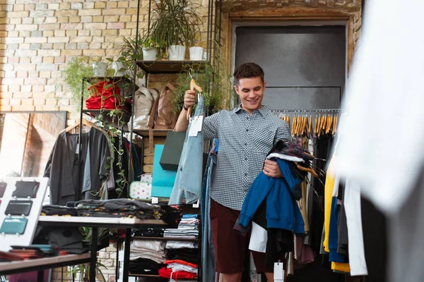 Happy man holding many clothes and smiling while visiting fashionable shop