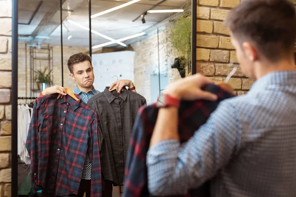 Tankevækkende ung mand ser i spejlet og ser på smukke skjorter - Stock-foto