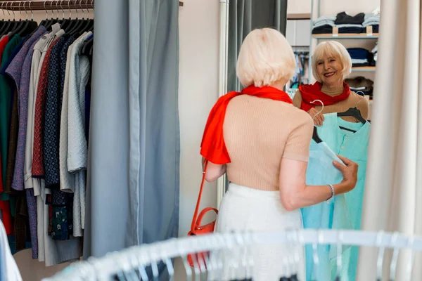 Φωτεινή ηλικιωμένη γυναίκα μπροστά από τον καθρέφτη των αγορών με ένα φόρεμα στα χέρια — Φωτογραφία Αρχείου