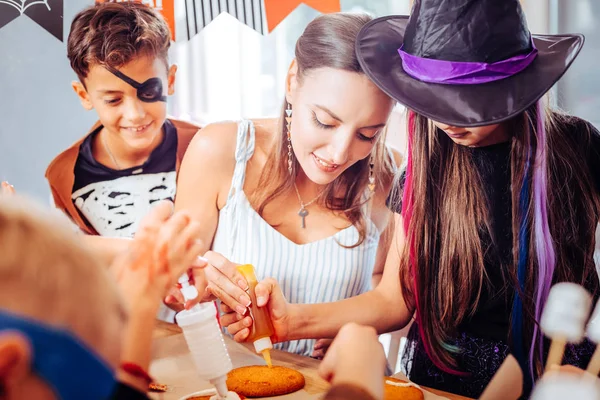 Caring mother helping her children with painted faces putting topping on cookies — Stock Photo, Image