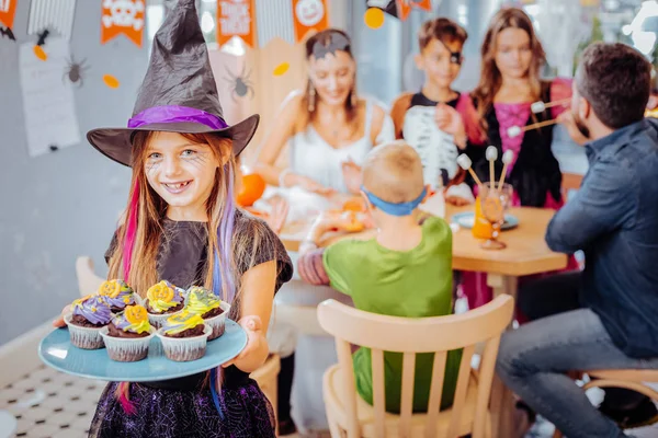 Smiling girl wearing Halloween wizard hat holding plate with celebration cupcakes