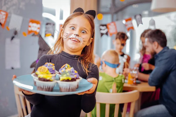 Funny girl wearing cat suit at the Halloween party holding plate with cupcakes