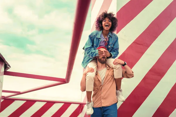 Laughing young woman closing eyes of boyfriend while sitting on his shoulders