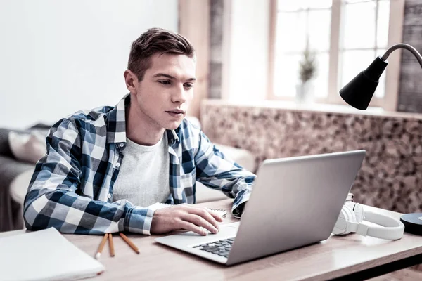 Спокойный студент смотрит на экран своего ноутбука, сидя за столом — стоковое фото
