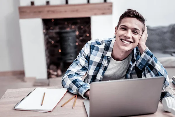 Positive student leaning his head on his hand and smiling while using his laptop — Stock Photo, Image