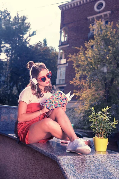 Chica brillante y elegante escuchando música en los auriculares y la lectura de libro interesante —  Fotos de Stock