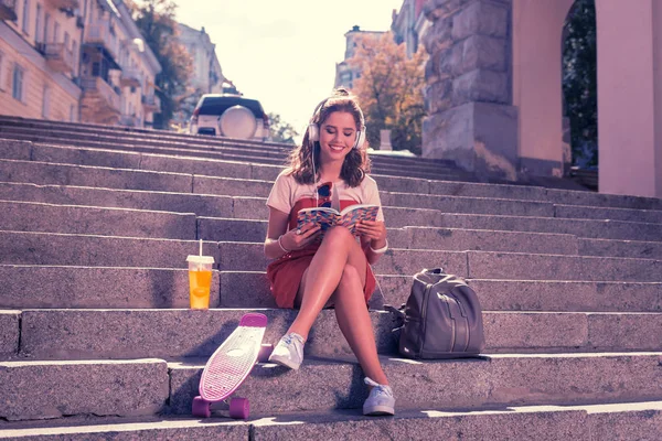 Söta leende flicka sitter på trappan nära hennes skateboard medan du läser bok — Stockfoto
