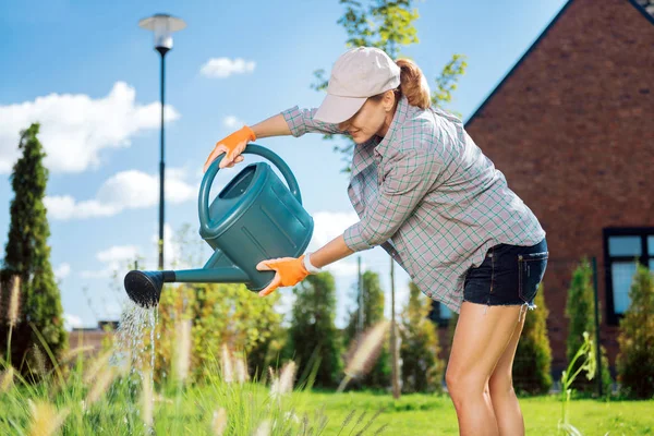 Blonďatá žena nosí ploché víčko nalévání vody na její rostliny mimo — Stock fotografie