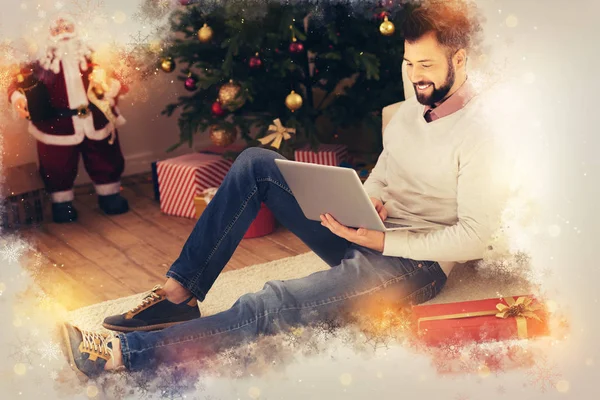 Knappe stijlvolle freelancer zittend op de vloer in de buurt van de kerstboom met zijn laptop — Stockfoto