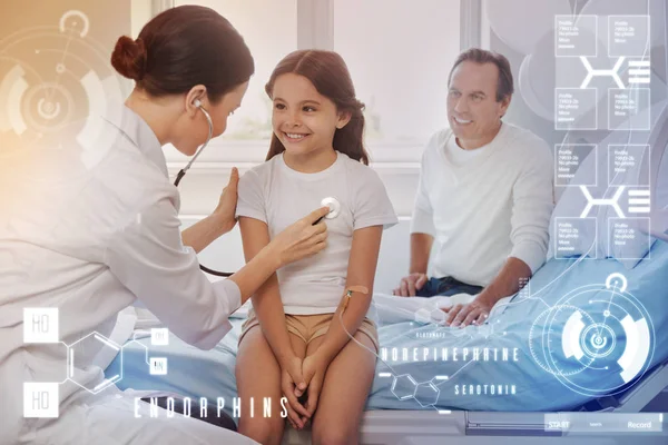 Chica sonriente sentada en la cama mientras un médico la examina con un estetoscopio — Foto de Stock