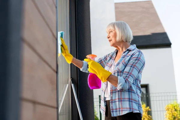 Señora mayor con guantes de color amarillo brillante sensación de ocupado lavando ventanas — Foto de Stock