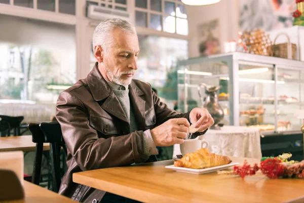 Homem de cabelos grisalhos sentado na padaria francesa comendo croissant e bebendo café — Fotografia de Stock