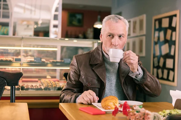 Dunkeläugiger älterer Mann trinkt morgens Kaffee in französischer Bäckerei — Stockfoto