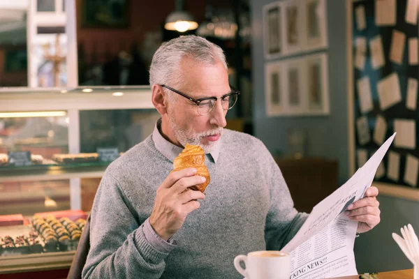 Бородатий чоловік в окулярах їсть круасан і читає новини — стокове фото