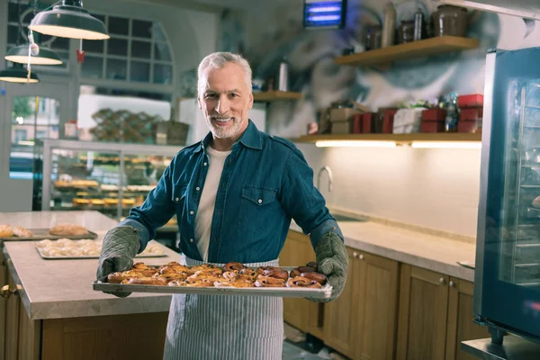 Beaming grey-haired baker holding whole tray with nice cinnamon buns