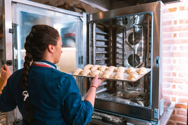 Baker dengan kepang tulang ikan menempatkan nampan dengan croissant ke dalam oven — Stok Foto