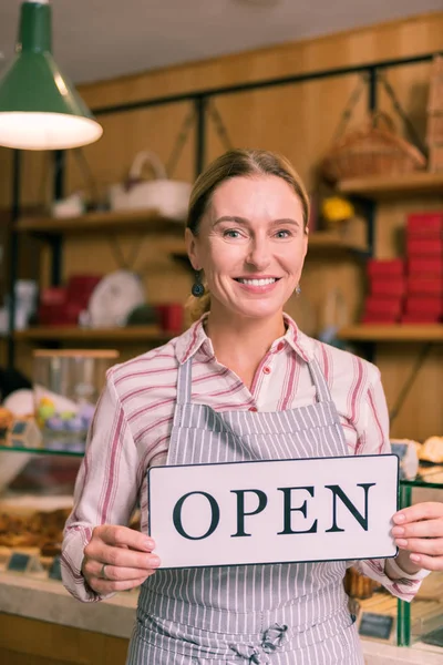 Smiling cheerful businesswoman wearing striped apron opening bakery