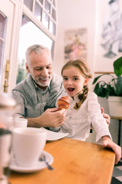 Cute stylish granddaughter wearing white shirt trying croissant — Stock Photo, Image