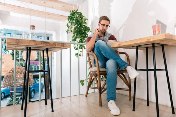 Homem elegante vestindo jeans e tênis brancos lendo livro na cafetaria — Fotografia de Stock