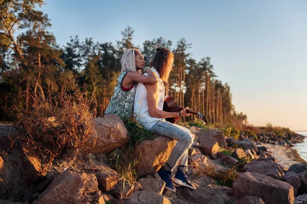 Жінка з білими дредлоками обіймає хлопця, сидячи на каменях з гітарою — стокове фото