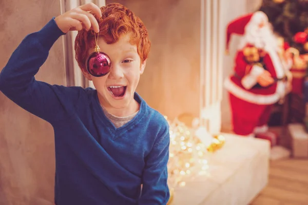 Joyful menino ruivo cobrindo seu olho com uma bugiganga de Natal — Fotografia de Stock