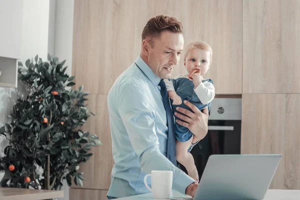 Hombre responsable ocupado usando el portátil y sosteniendo a un bebé . —  Fotos de Stock