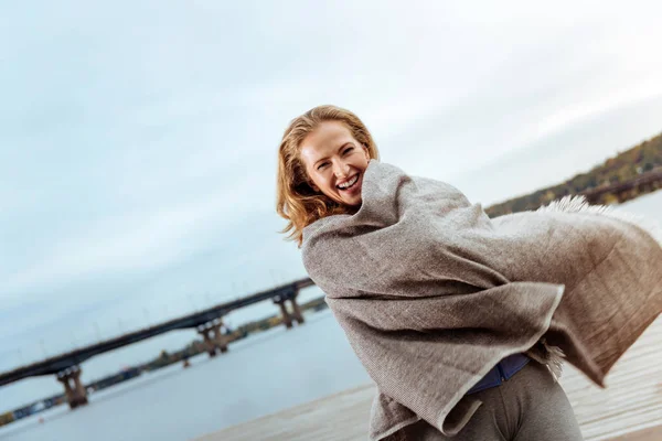 Glad ung kvinna som bär en pläd nära vattnet — Stockfoto