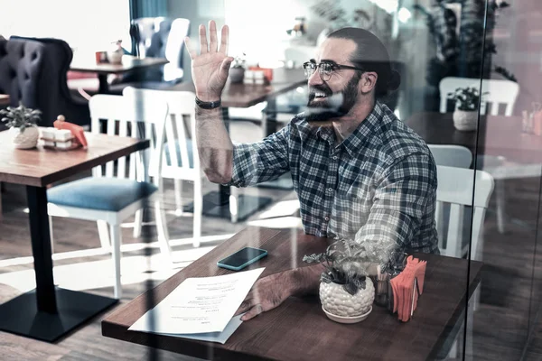 Pleasant bebaarde man glimlachen en zwaaien van zijn hand bijeenkomst vriend in cafetaria — Stockfoto