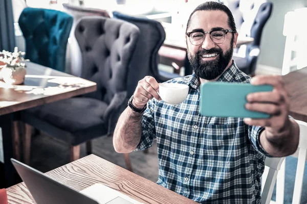 Beaming bearded man feeling relaxed making selfie and drinking coffee — Stock Photo, Image