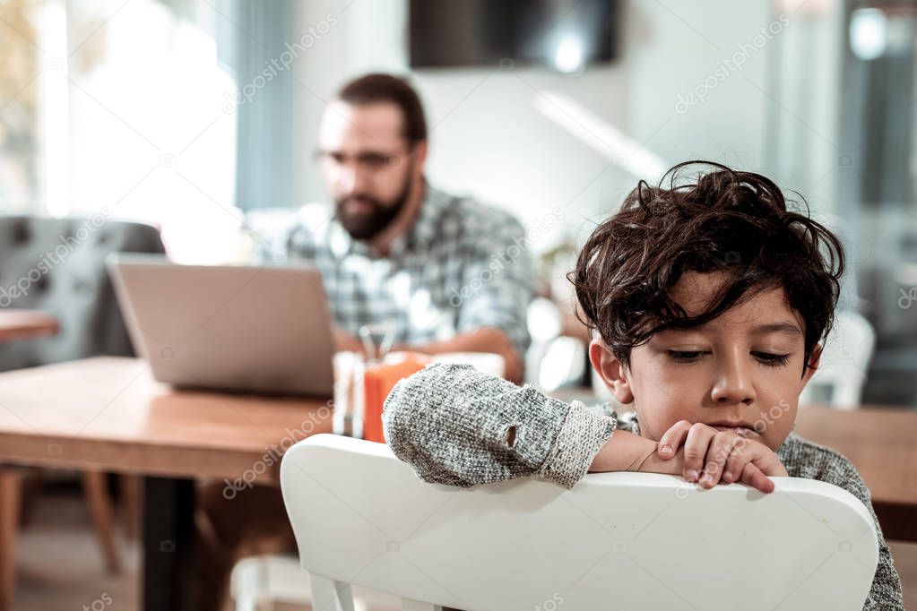 Cute dark-haired son feeling bored while father working on laptop