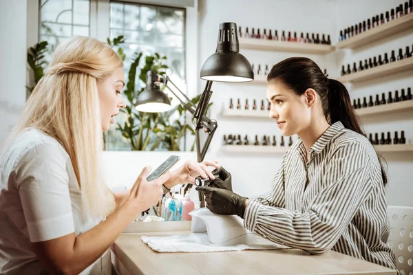Blonde-haired woman holding her smartphone sitting in front of nail artist — Stock Photo, Image
