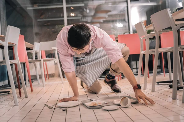Clean floor. Young dark-haired waiter cleaning the floor after dropping tray with food and coffee