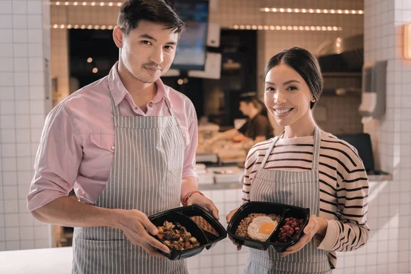 Dos camareros radiantes agradables sosteniendo cajas de almuerzo agradables con comida sabrosa — Foto de Stock
