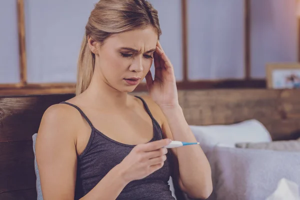 Fair-haired young woman having fever and checking thermometer — Stock Photo, Image