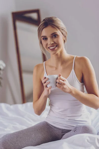 Fröhliche junge Frau sitzt auf dem Bett und trinkt Kaffee — Stockfoto