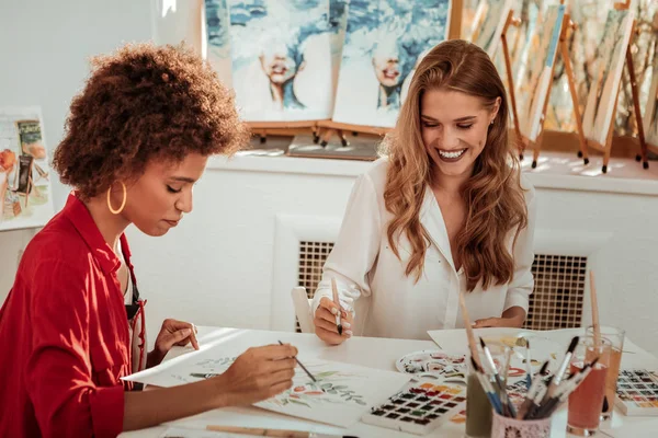 Beaming blonde-haired artist smiling broadly painting with friend — Stock Photo, Image