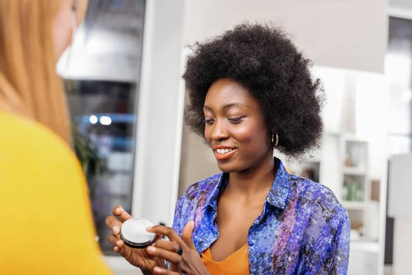 Schöne dunkelhäutige Stylistin mit lockigem Haar, die ihrem Kunden Puder zeigt — Stockfoto