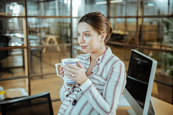 Pregnant appealing woman drinking tasty warm tea in office