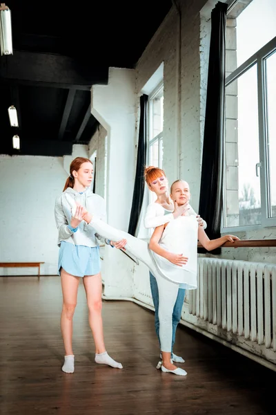 Adolescente rubia de pelo largo con ropa deportiva blanca haciendo piernas estiradas cerca de la barra de ballet — Foto de Stock